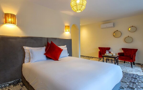A hotel room with a large bed, two red chairs, a small table, and a decorative wall hanging.