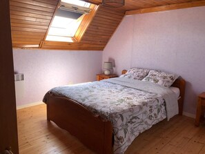 Quarto com teto inclinado, claraboia, cama de madeira, mesa de cabeceira com abajur e aquecedor fixado na parede.