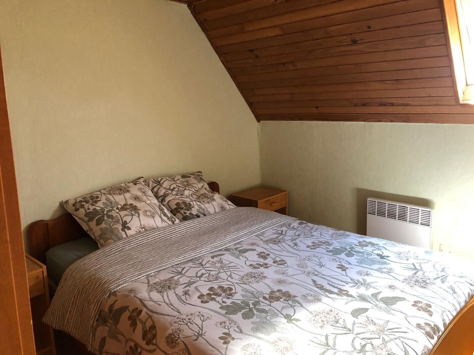 Une chambre avec un plafond en pente, un lit avec une literie à motifs floraux, une table de chevet et un climatiseur fixé au mur.