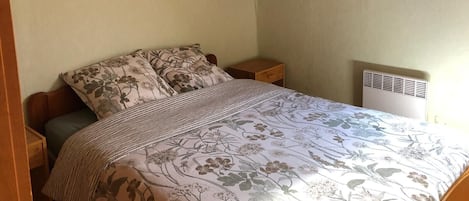 Une chambre avec un plafond en pente, un lit avec une literie à motifs floraux, une table de chevet et un climatiseur fixé au mur.