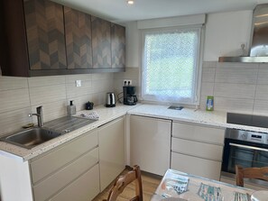 A modern kitchen with a built-in dishwasher, a coffee maker, and a window with lace curtains. - Holiday Home 'Maison De Vacances Crozon Jardin' with Sea View, Private Terrace and Wi-Fi (Crozon)