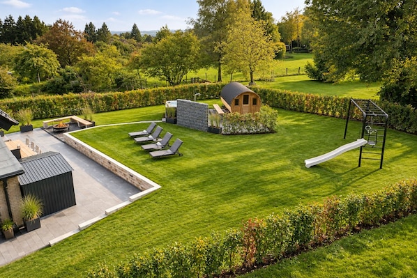 A well-maintained garden with a wooden playhouse, a swing set, and a seating area with lounge chairs.