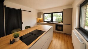 A modern kitchen with a central island, a built-in oven, and a window providing a view of the outside. - Vennchalet - Vacation Home for 8 People with a Sauna and Hot Tub (Monschau)