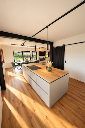 A modern kitchen with a large island, wooden flooring, and a view of the outdoors.