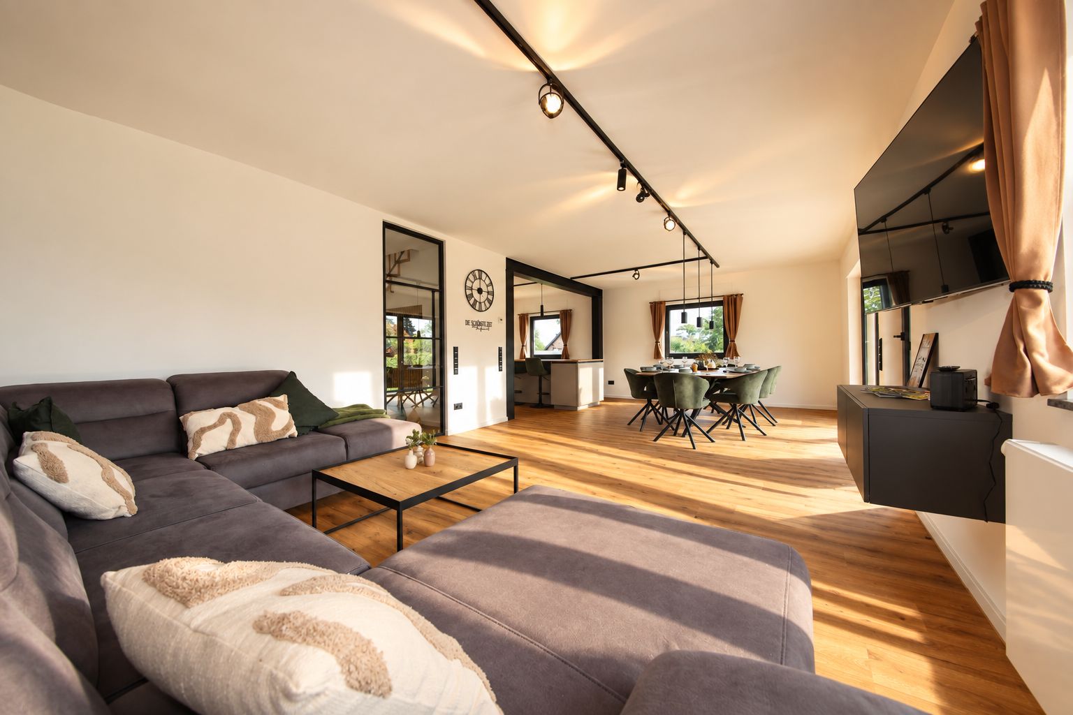 Ein modernes Wohnzimmer mit einer Eckcouch, einem Essbereich mit Tisch und Stühlen sowie einer Küchenzeile und einem Fernseher.