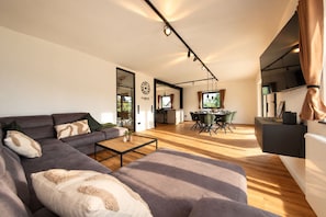 A modern living room with a sectional sofa, a dining area with a table and chairs, and a kitchen area with a TV.