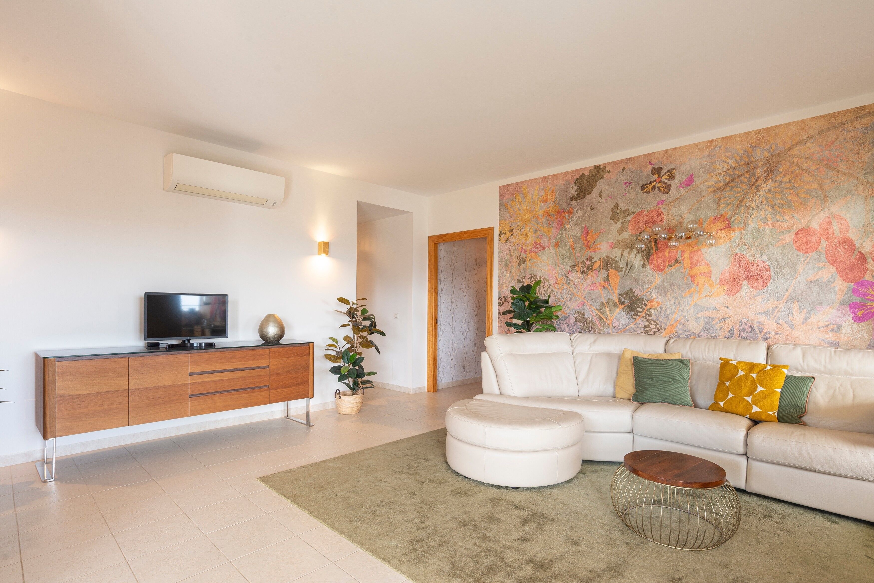 Una sala moderna con un sofá blanco, un televisor sobre un mueble de madera, una mesa de centro redonda y un cuadro abstracto colorido en la pared.