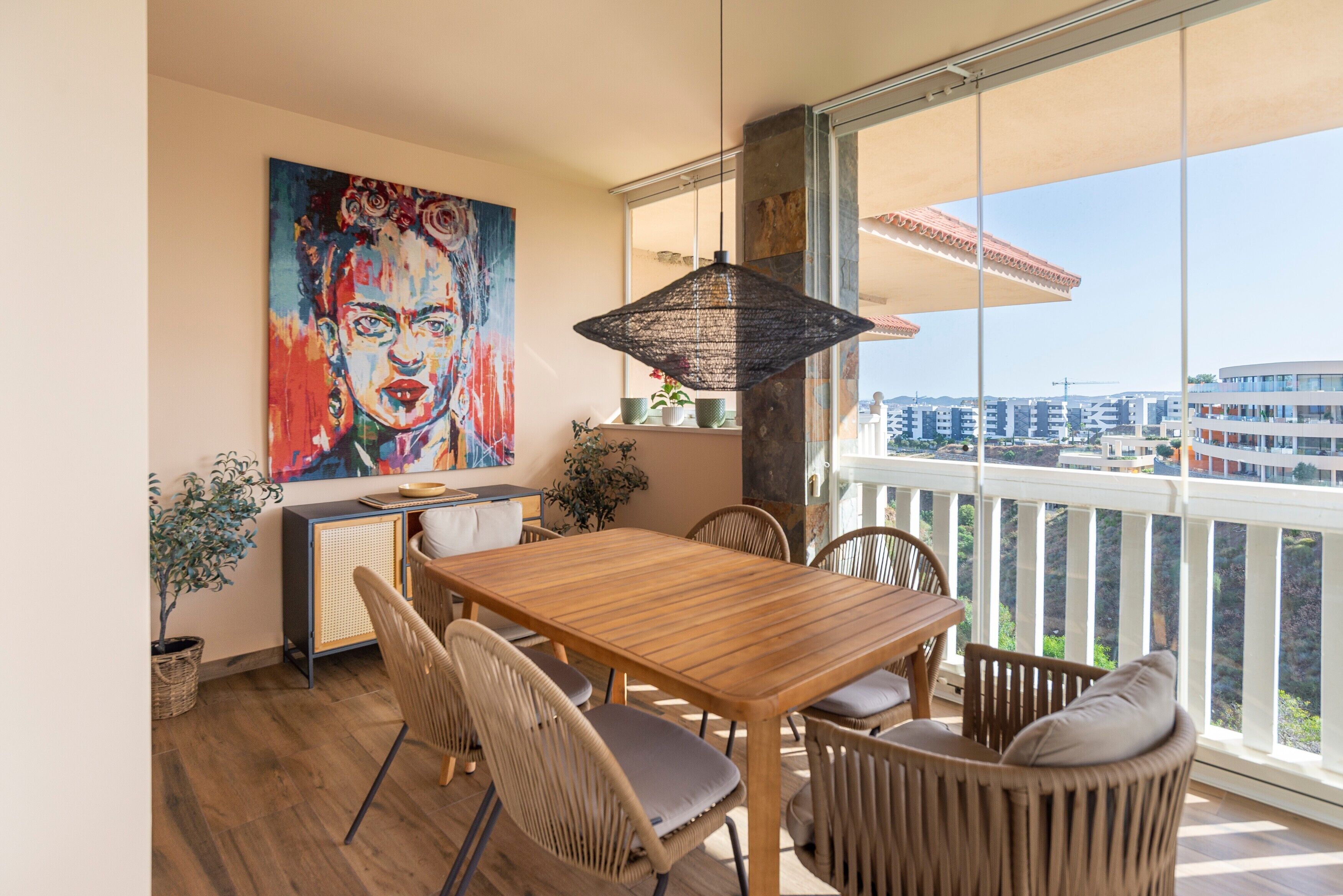 Un coin repas avec une table en bois et des chaises en osier, une grande fenêtre donnant sur des immeubles et un tableau coloré accroché au mur.