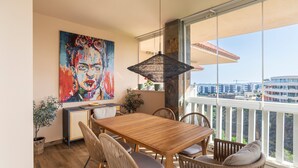 A dining area with a wooden table and wicker chairs, a large window with a view of buildings, and a colorful painting on the wall. - Holiday Home 'Cozy 3BR Apartment Higuerón' with Mountain View, Wi-Fi and Air Conditioning (Benalmádena)