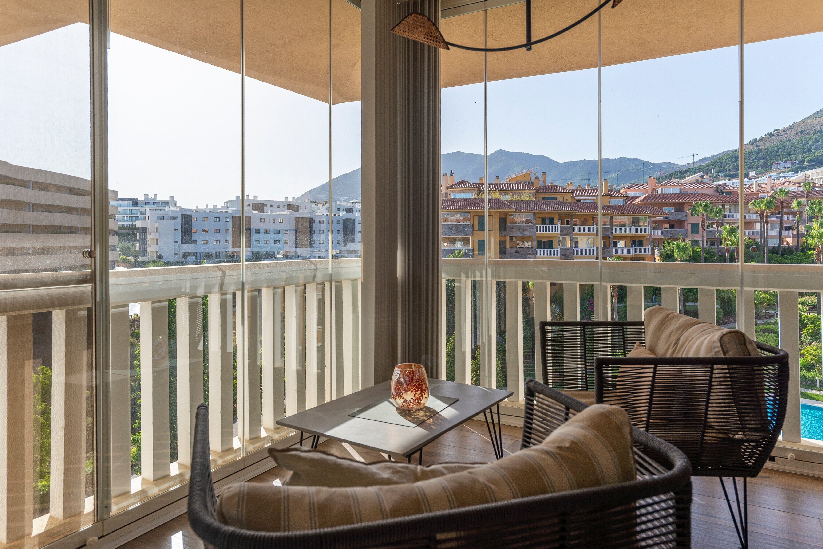 Un balcon agrémenté de meubles en osier, d’une table en verre et offrant une vue sur les bâtiments et les montagnes.