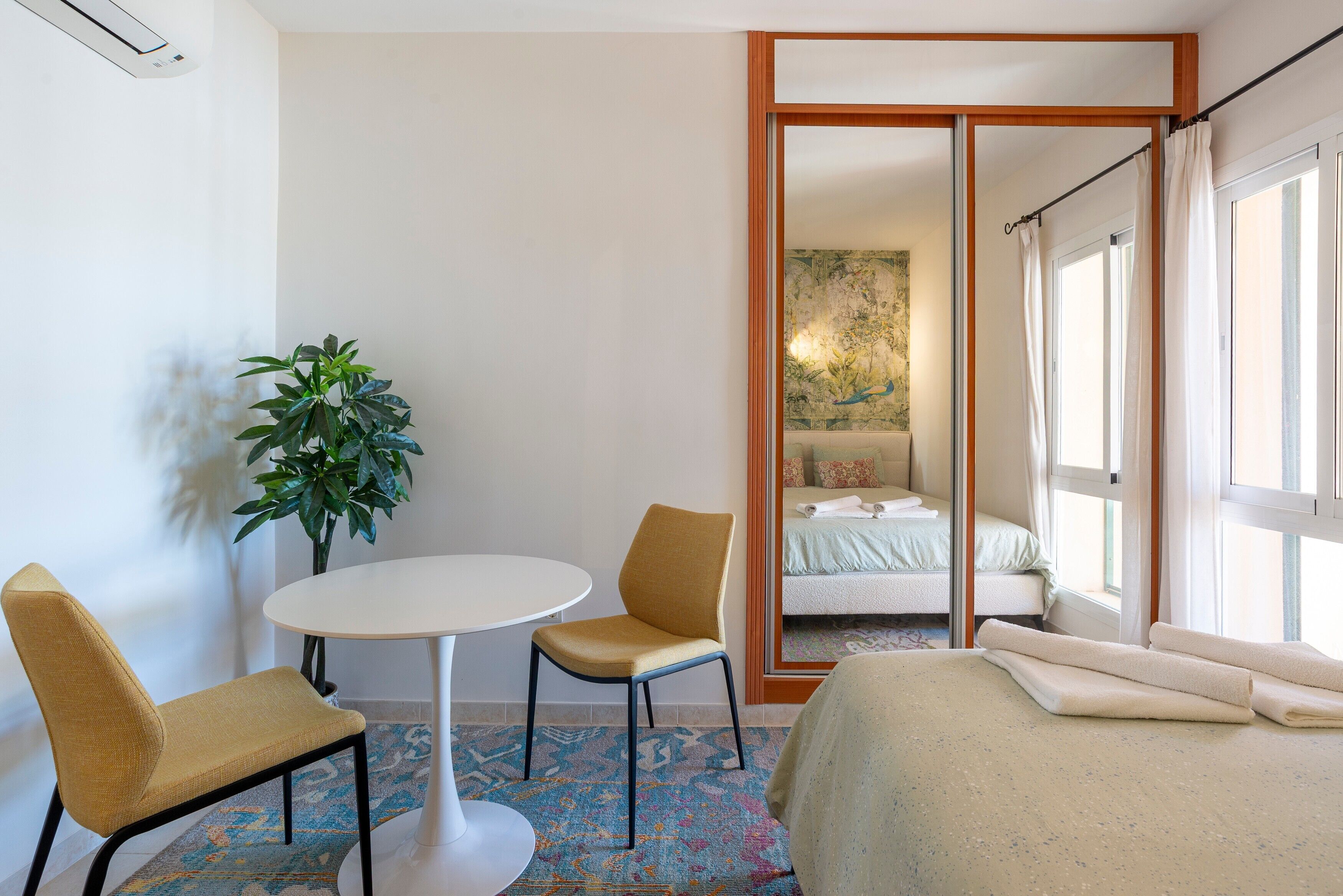 Une chambre d’hôtel avec un lit, une petite table, deux chaises, une plante en pot et un grand miroir qui reflète la pièce.