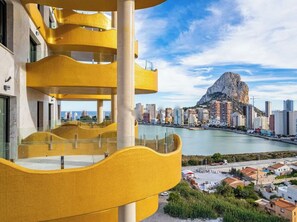 A modern building with a yellow facade and balconies overlooking a coastal cityscape with a prominent rock formation. - Luxury 3-bedroom apartment with panoramic views. (Calpe)