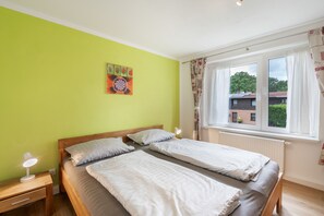 A room with two beds, a nightstand with a lamp, a window with curtains, and a painting on the wall.