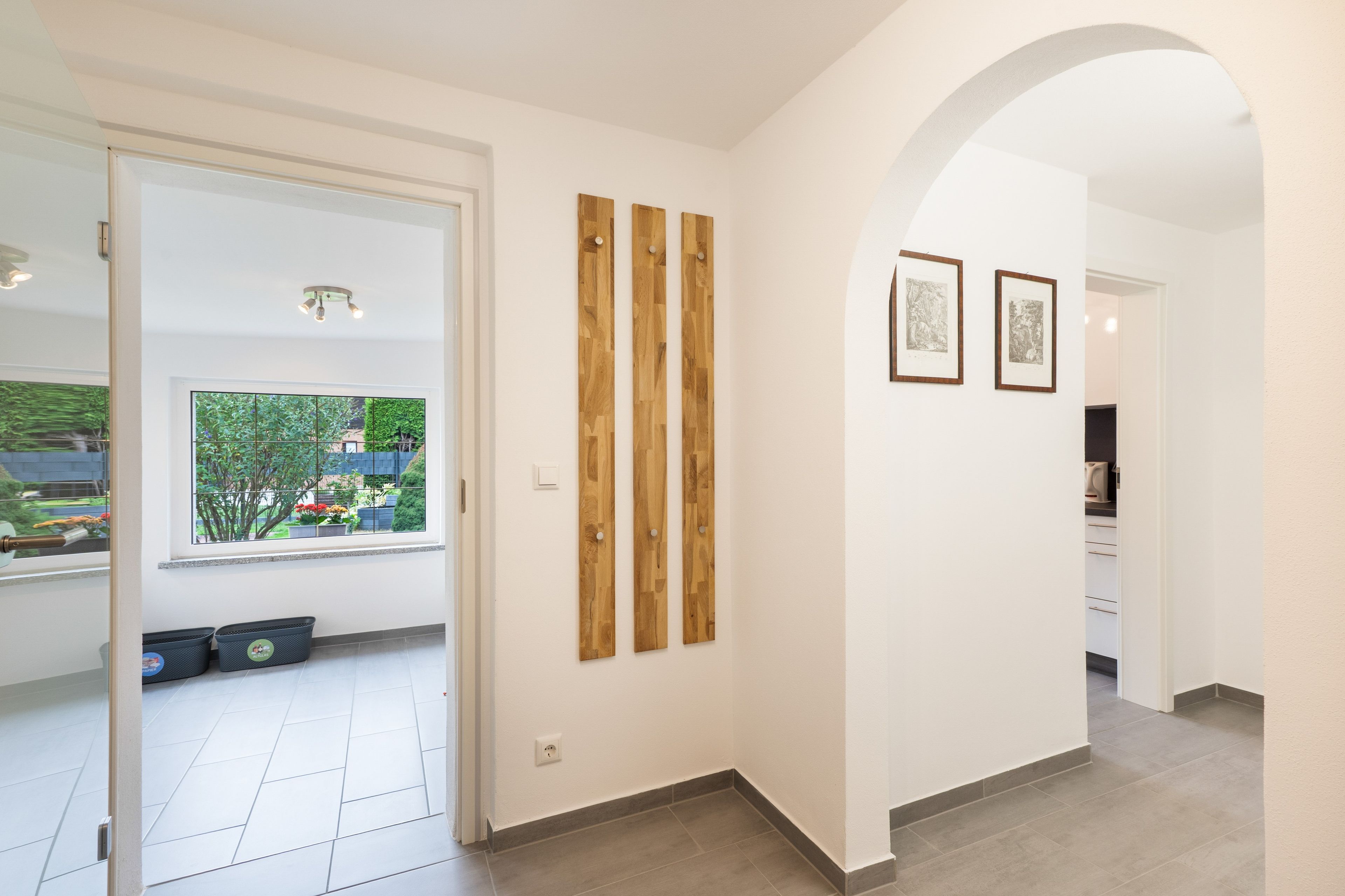 Ein Flur mit einer weißen Tür, die zu einem Zimmer mit Fenster und Gartenblick führt. An der Wand befinden sich zwei Holzpaneele und zwei gerahmte Bilder.