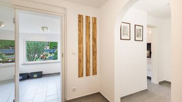 Ein Flur mit einer weißen Tür, die zu einem Zimmer mit Fenster und Gartenblick führt. An der Wand befinden sich zwei Holzpaneele und zwei gerahmte Bilder.