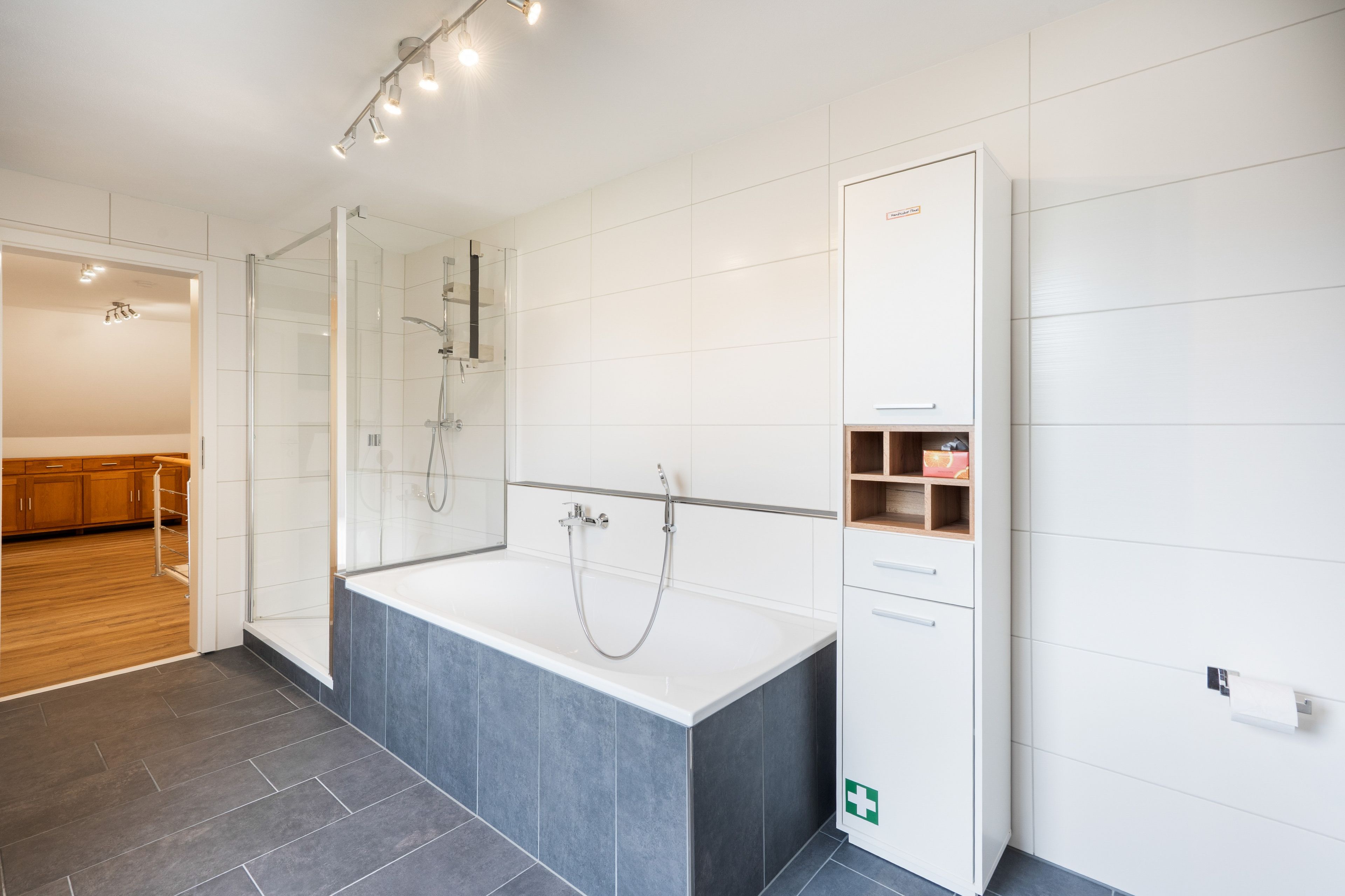 Ein modernes Badezimmer mit Duschkabine aus Glas, einer freistehenden Badewanne und einem weißen Schrank mit einem grünen Notausgangsschild.