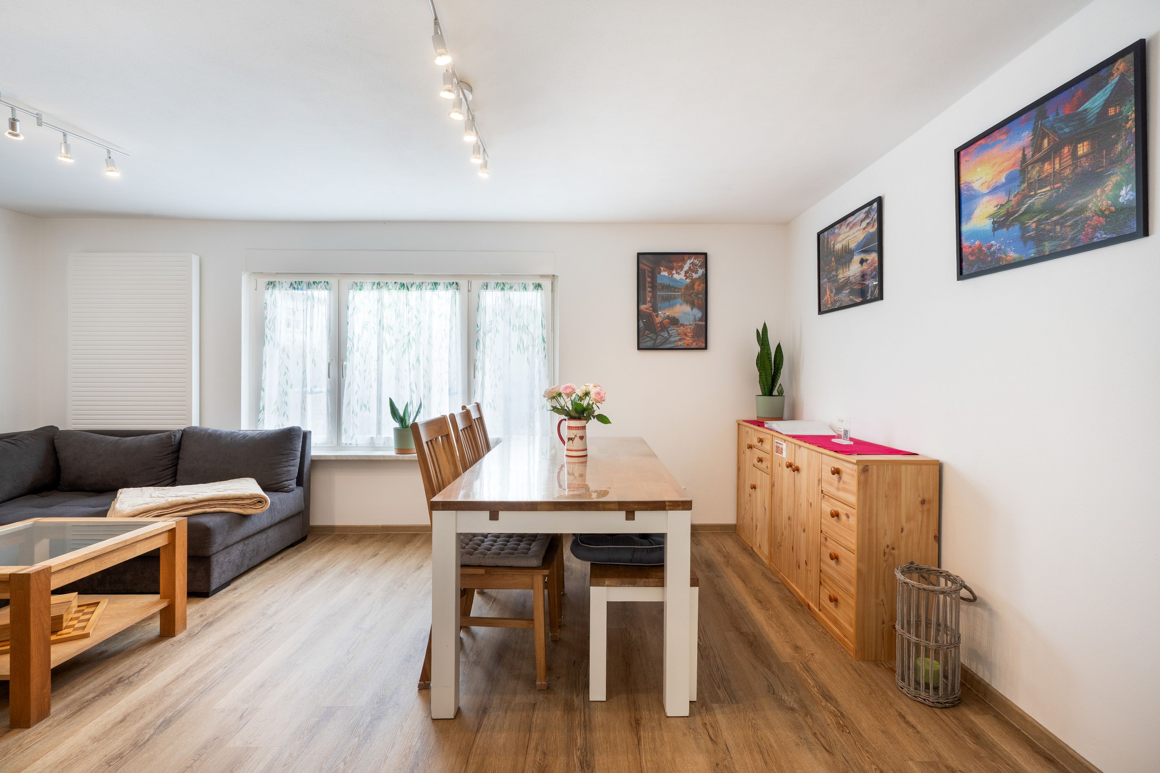 Un salon moderne avec un canapé, une table à manger et des chaises. Il y a une fenêtre avec des rideaux, une plante et des œuvres d’art accrochées au mur.