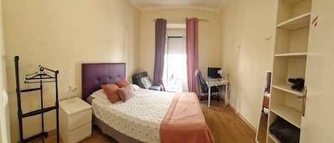 A bedroom with a bed, a desk, a chair, a wardrobe, and a hanging clothes rack.