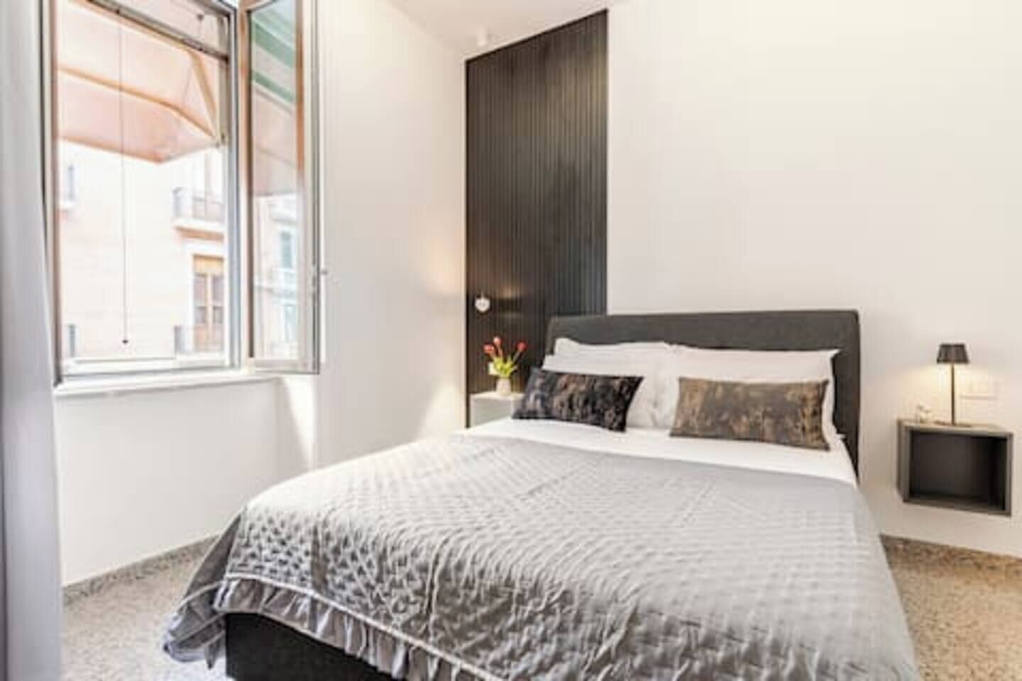 A bedroom with a bed, a nightstand, a lamp, and a window with a view of buildings.