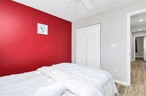 Un dormitorio con pared roja, cama blanca y puerta que da a otra habitación.