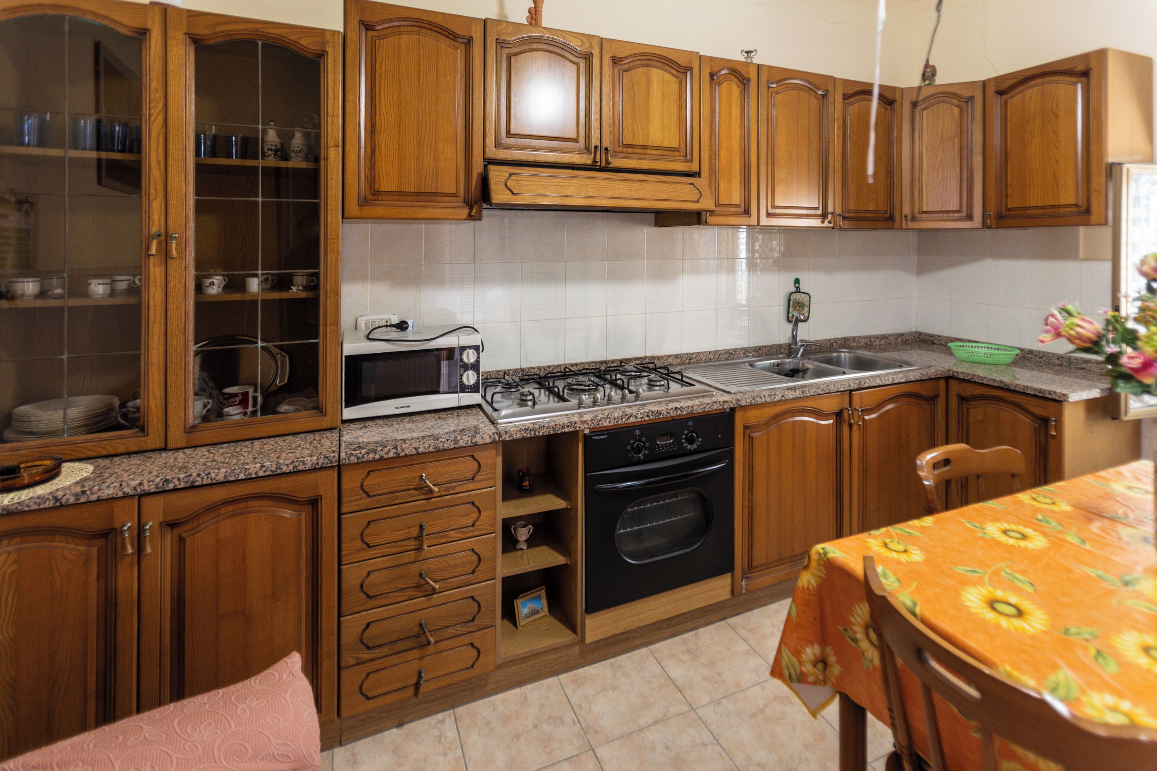 Une cuisine avec des meubles en bois, une cuisinière, un four et une table à manger recouverte d’une nappe  à motifs floraux.