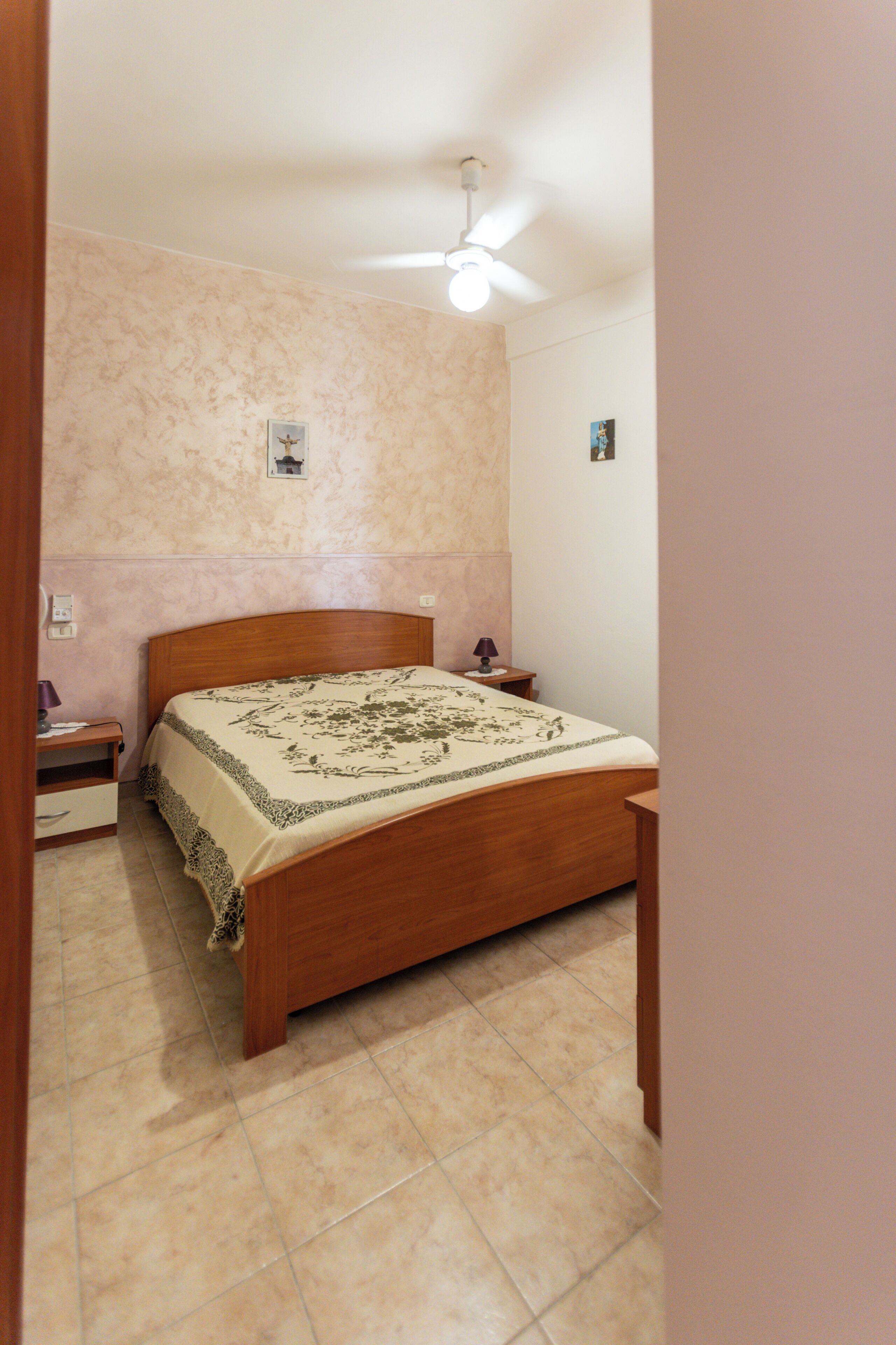 Une chambre à coucher avec un lit en bois, une table de chevet, une lampe, un ventilateur de plafond et un interrupteur fixé au mur.