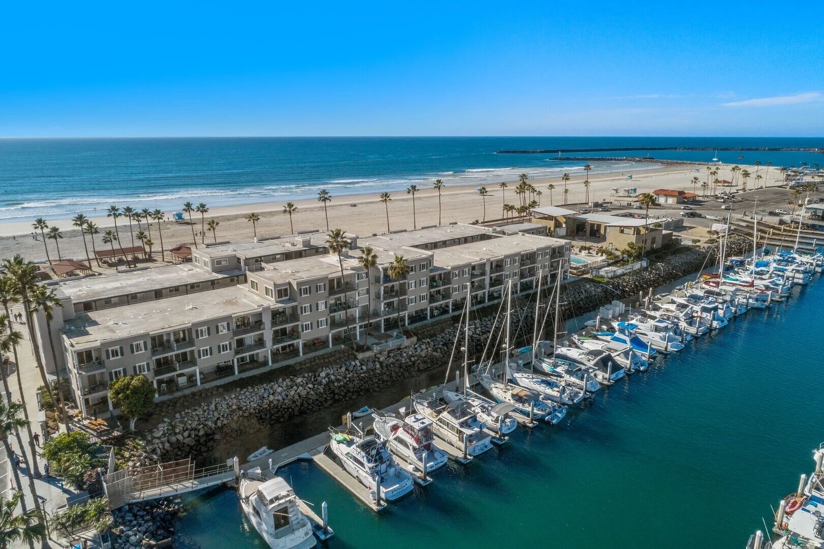 A coastal hotel with multiple floors, a marina with boats, and a beach in the background.