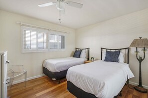 A bedroom with two beds, a ceiling fan, a window with blinds, a bedside lamp, and a clear chair. - Beachfront 2BR Harbor Condo w/ Ocean View Balcony (Oceanside)