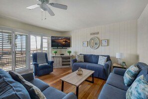 A living room with a sofa, armchair, and coffee table. There is a television mounted on the wall, and a ceiling fan is visible. - Beachfront 2BR Harbor Condo w/ Ocean View Balcony (Oceanside)