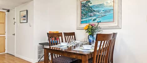 Un comedor con mesa de madera dispuesta para cuatro personas, sillas, un florero con flores y un cuadro enmarcado en la pared.