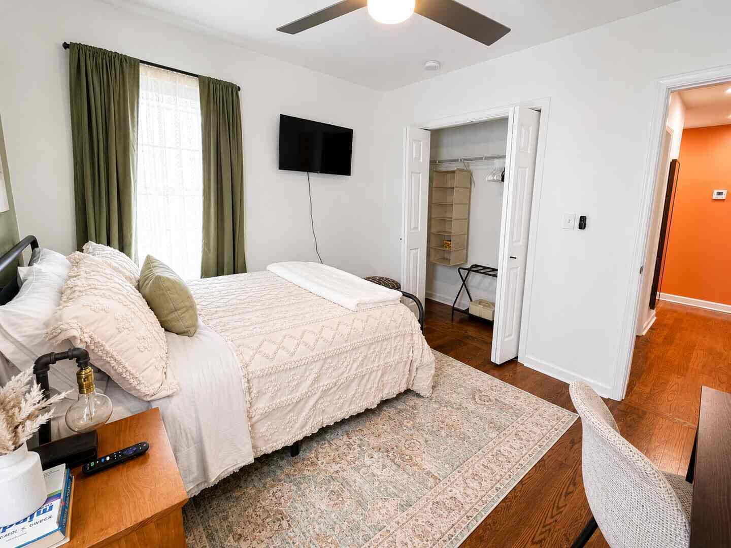 Een slaapkamer met een bed, een televisie, een bureau met stoel, een raam met gordijnen en een deur naar een andere kamer.