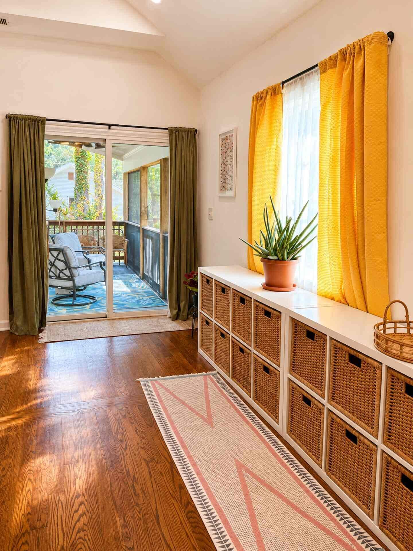 Una habitación con piso de madera, un vidrio corredizo que da a un espacio al aire libre, una mesa consola blanca con cestas y una planta en maceta.
