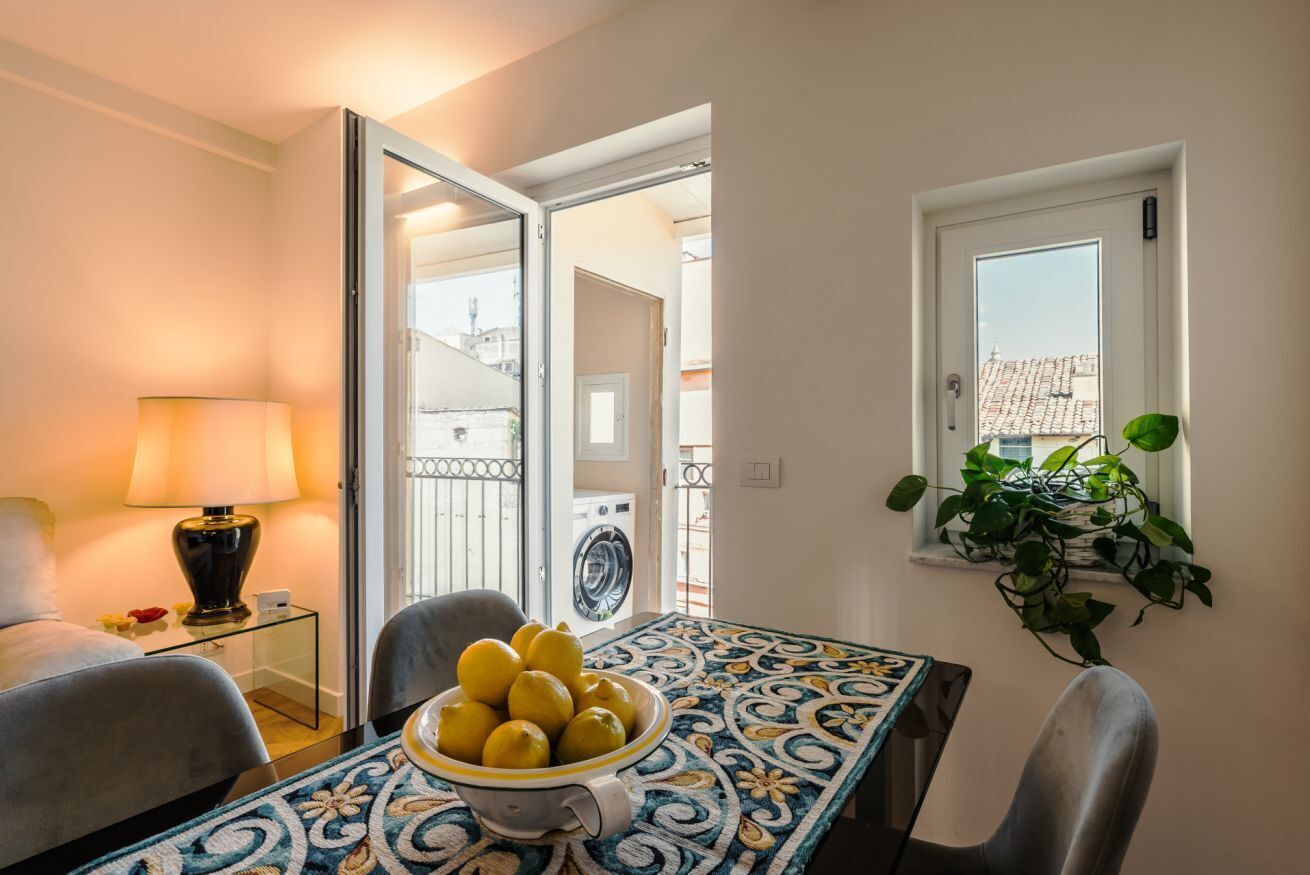 A room with a dining table, chairs, a lamp, a bowl of lemons, a washing machine, and a potted plant.