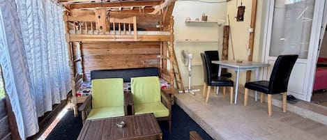 A cozy cabin-style room with a wooden ceiling, a bed with a headboard, two green chairs, a small table, a dining table with chairs, and a fan.
