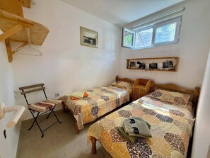 Un dormitorio con dos camas, una mesita, una silla, una ventana y una estantería con fotografías.