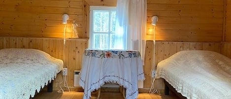 A cozy cabin room with two beds, a window with curtains, a table with a floral tablecloth, and two wall-mounted lamps.