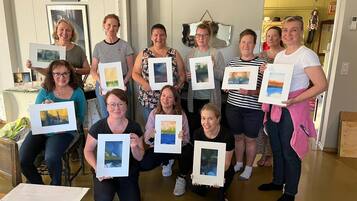 A group of people holding framed artworks in an indoor setting with a wooden floor and a door in the background.