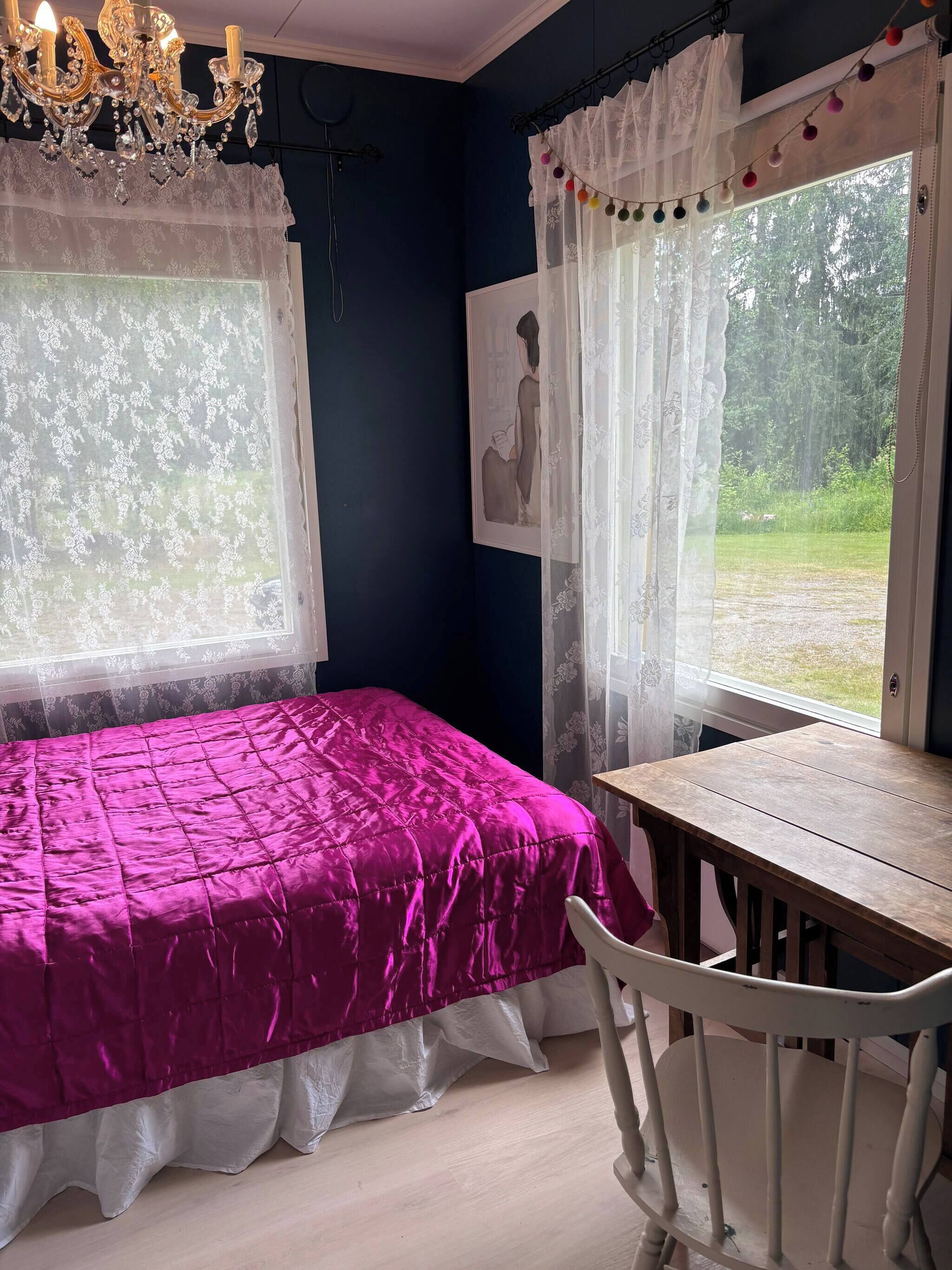 Ein Zimmer mit einem Bett, einem Tisch und einem Stuhl. Zwei Fenster mit Vorhängen, an einer Wand hängt ein Bild.