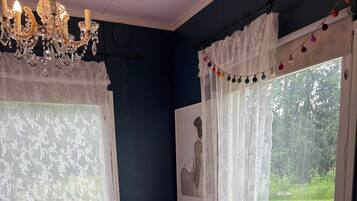 Ein Zimmer mit einem Bett, einem Tisch und einem Stuhl. Zwei Fenster mit Vorhängen, an einer Wand hängt ein Bild.