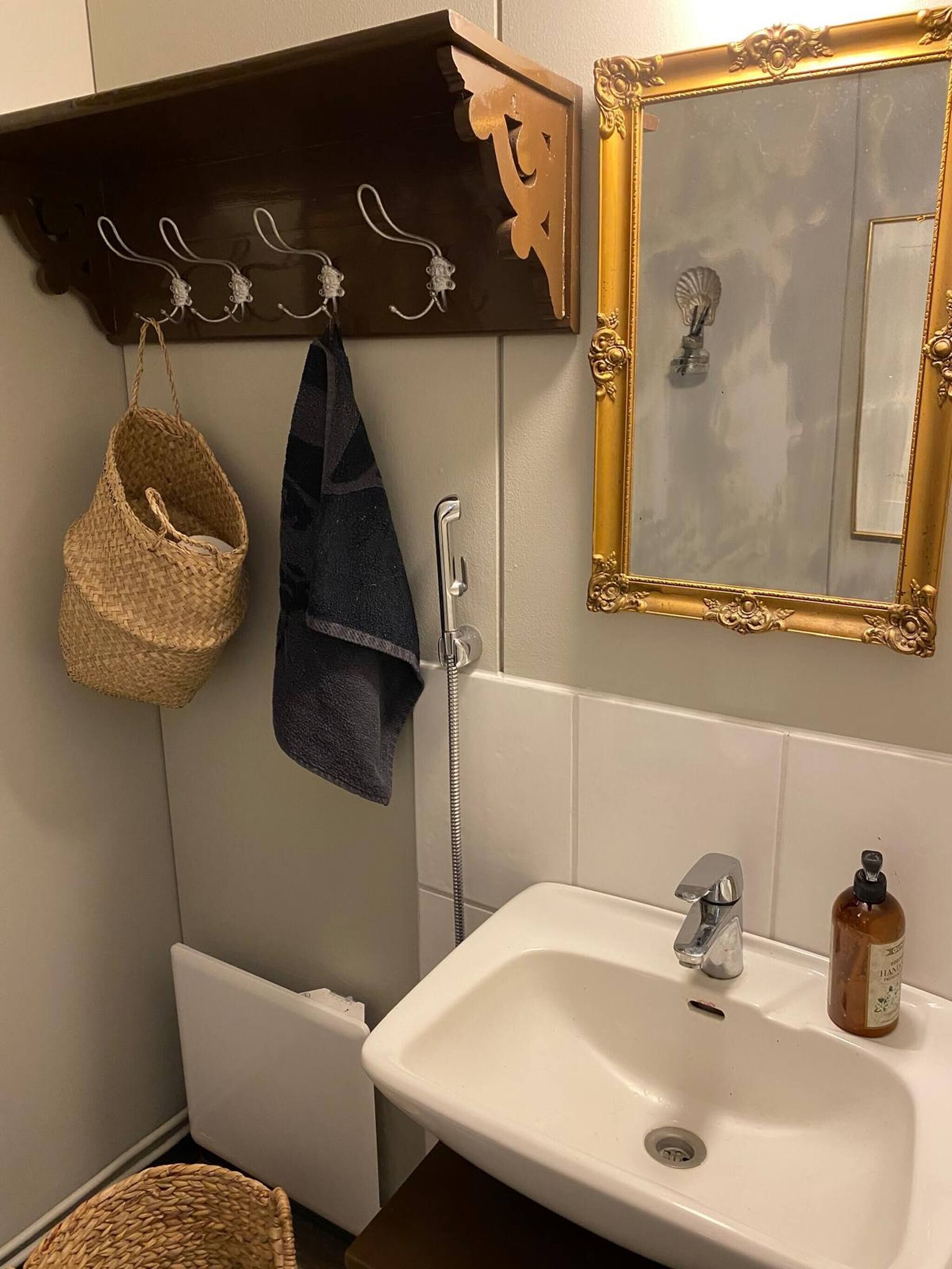 A bathroom with a white sink, a gold-framed mirror, a towel rack with a woven basket and a dark towel, and a shower head.