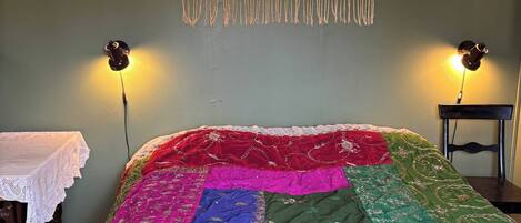 Un lit recouvert d’une courtepointe aux couleurs vives, une tapisserie murale et deux lampes fixées au mur.