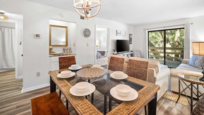 A modern living room with a glass dining table, wicker furniture, a large window with a view, and a flat-screen TV. - Fiddlers Cove Gated Resort- Pools- Hot Tub - Tennis - Pickle Ball walk to Beach! (Hilton Head Island)