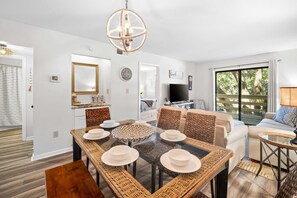 A modern living room with a glass dining table, wicker furniture, a large window with a view, and a flat-screen TV. - Fiddlers Cove Gated Resort- Pools- Hot Tub - Tennis - Pickle Ball walk to Beach! (Hilton Head Island)