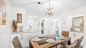 A modern kitchen with white cabinetry, a central island with a glass top, and a dining area with wicker chairs and a glass table. - Fiddlers Cove Gated Resort- Pools- Hot Tub - Tennis - Pickle Ball walk to Beach! (Hilton Head Island)