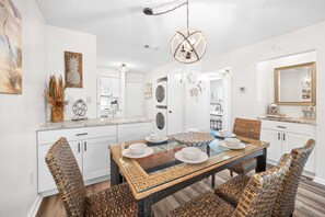 A modern kitchen with white cabinetry, a central island with a glass top, and a dining area with wicker chairs and a glass table. - Fiddlers Cove Gated Resort- Pools- Hot Tub - Tennis - Pickle Ball walk to Beach! (Hilton Head Island)
