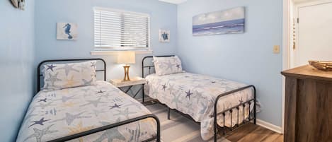 A bedroom with two beds, a window with blinds, a nightstand with a lamp, and a painting on the wall.
