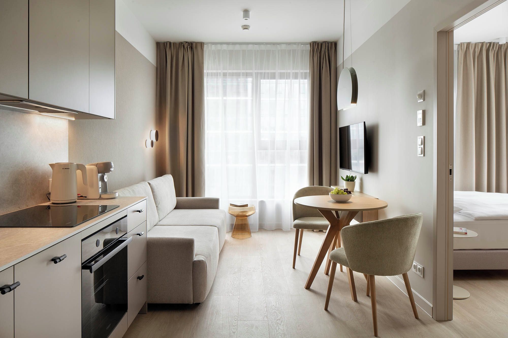 Ein modernes Hotelzimmer mit Küchenzeile, einer Couch, einem Esstisch mit Stühlen, einem an der Wand montierten Fernseher und einem großen Fenster mit Vorhängen.