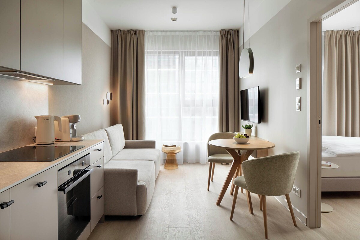 A modern hotel room with a kitchenette, a sofa, a dining table with chairs, a TV mounted on the wall, and a large window with curtains.