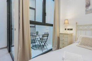A bedroom with a bed, a bedside table, a lamp, and a window with curtains. - Luxurious apartment with a patio in downtown Seville (Sevilla)