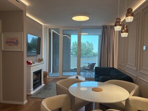 Un salon moderne avec une cheminée, une table ronde entourée de chaises, un téléviseur fixé au mur et un balcon agrémenté de mobilier d’extérieur.